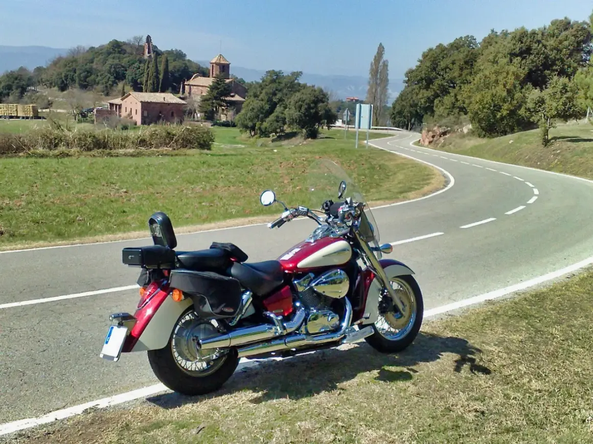 Honda VT 750 C Shadow 2007 ficha técnica. Moto custom roja y crema aparcada en una carretera rural con un paisaje de colinas y edificios antiguos.