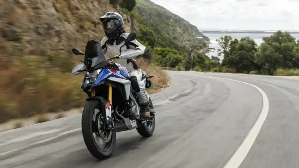 Motociclista en una maxi trail recorre una carretera sinuosa con vistas al mar y montañas verdes.