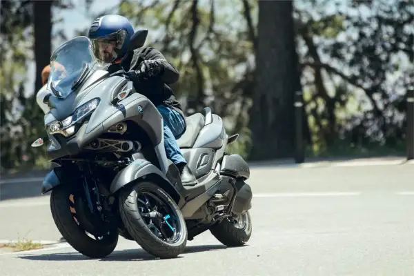 Hombre en casco azul conduce un triciclo Yamaha Tricity 500 por una carretera arbolada, inclinándose en una curva.